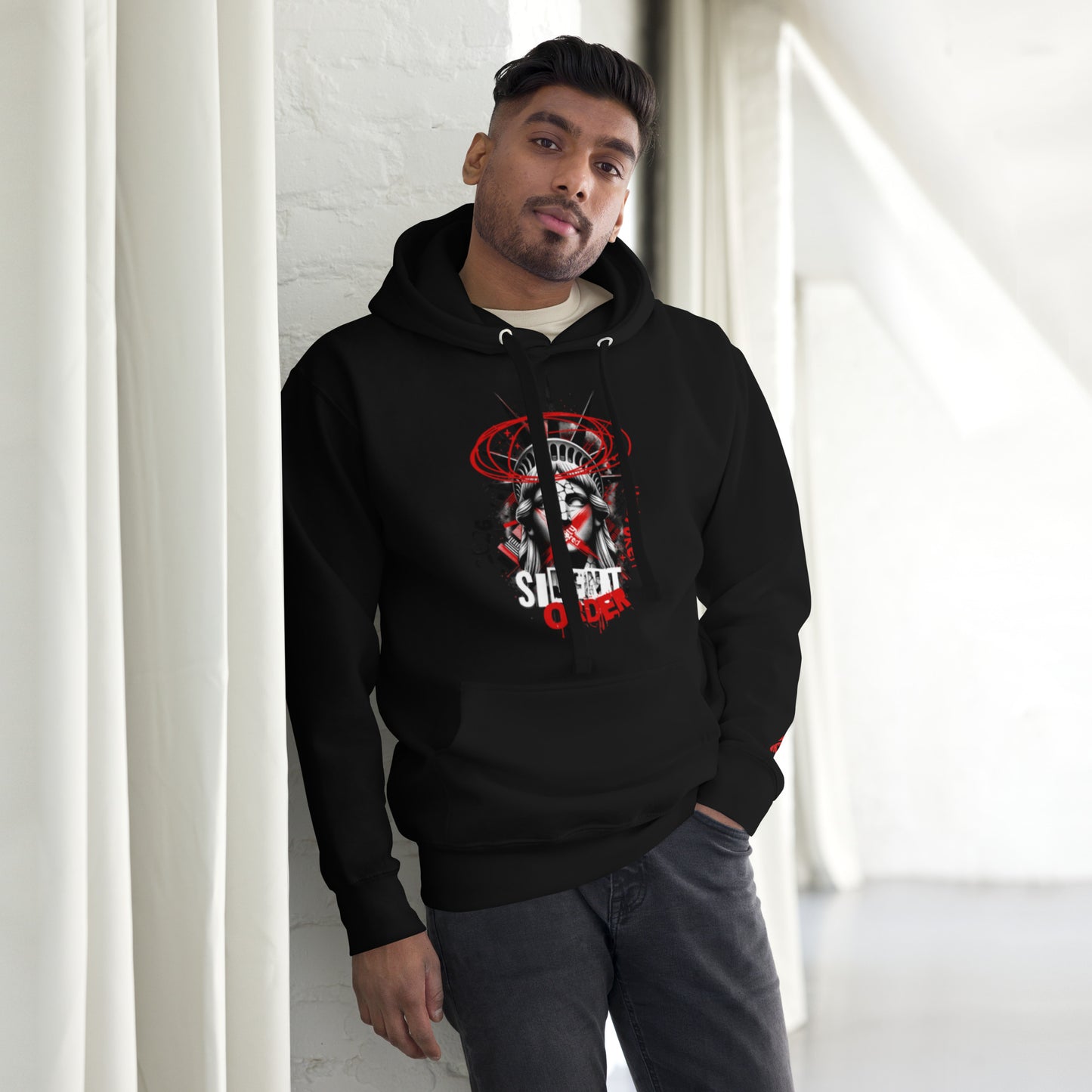 Man wearing a black hoodie with a graphic design, standing against a white wall.
