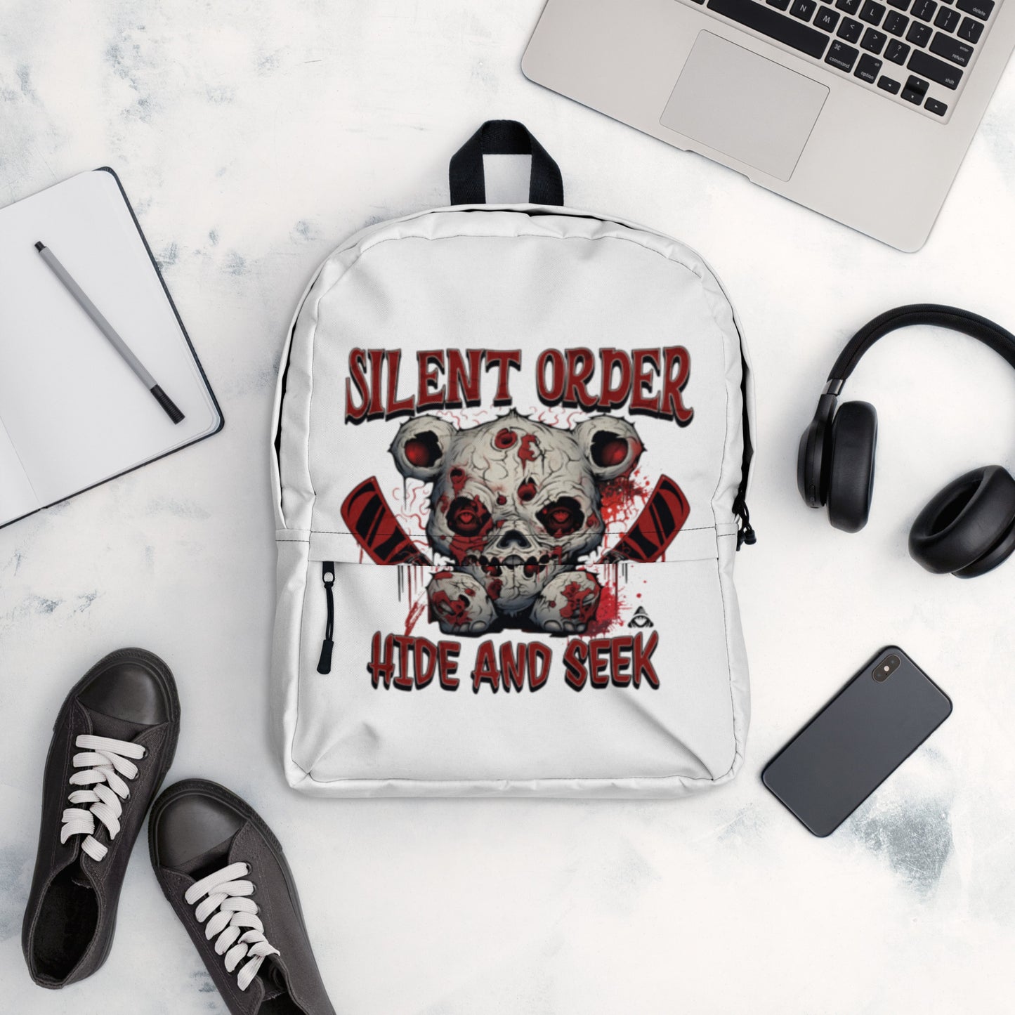 White backpack with graphic design on a marble surface with laptop, shoes, and phone.