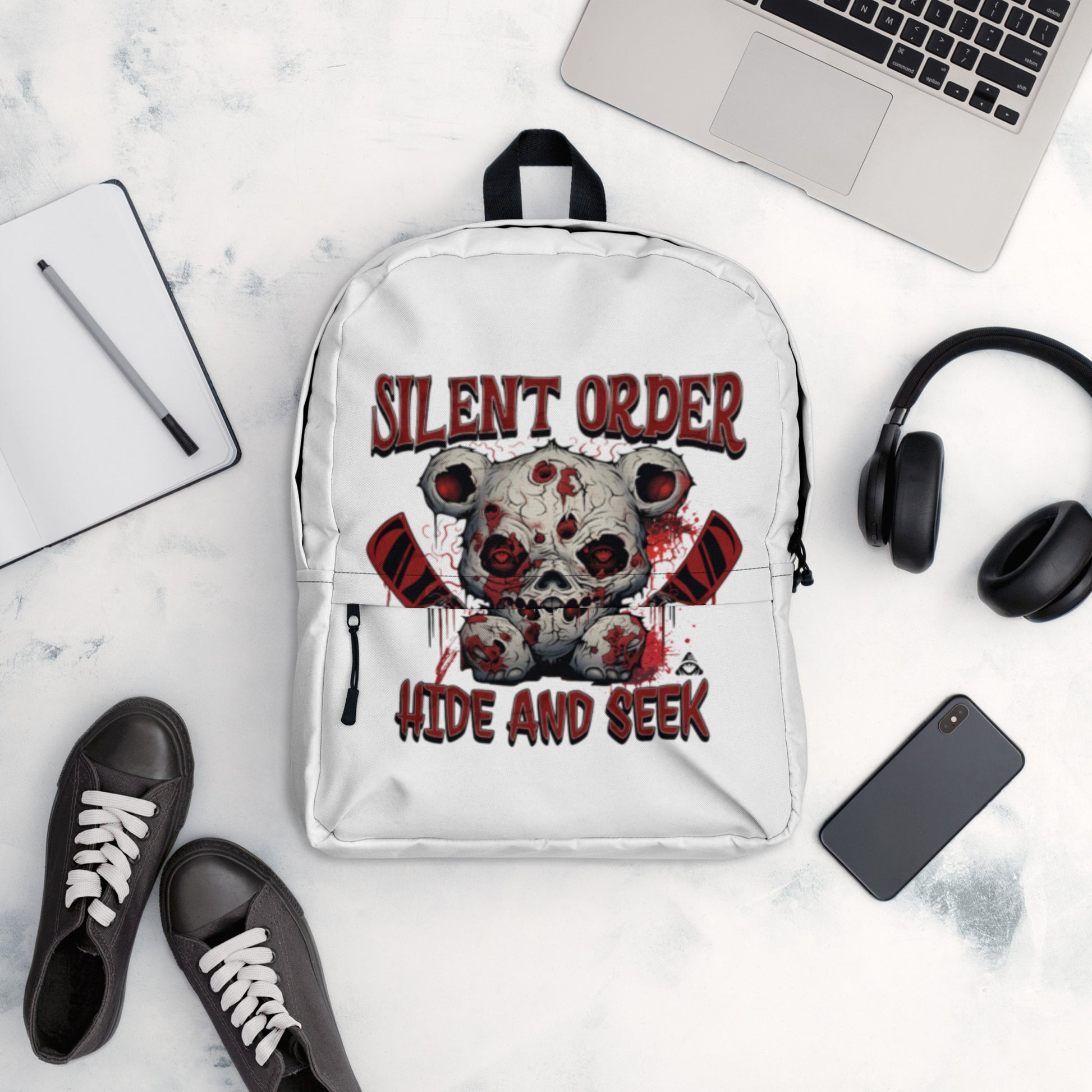 White backpack with graphic design on a marble surface with laptop, shoes, and phone.