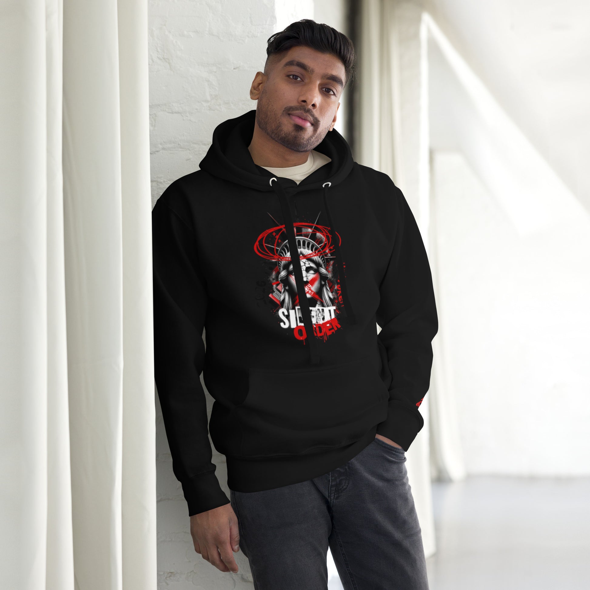 Man wearing a black hoodie with a graphic design, standing against a white wall.